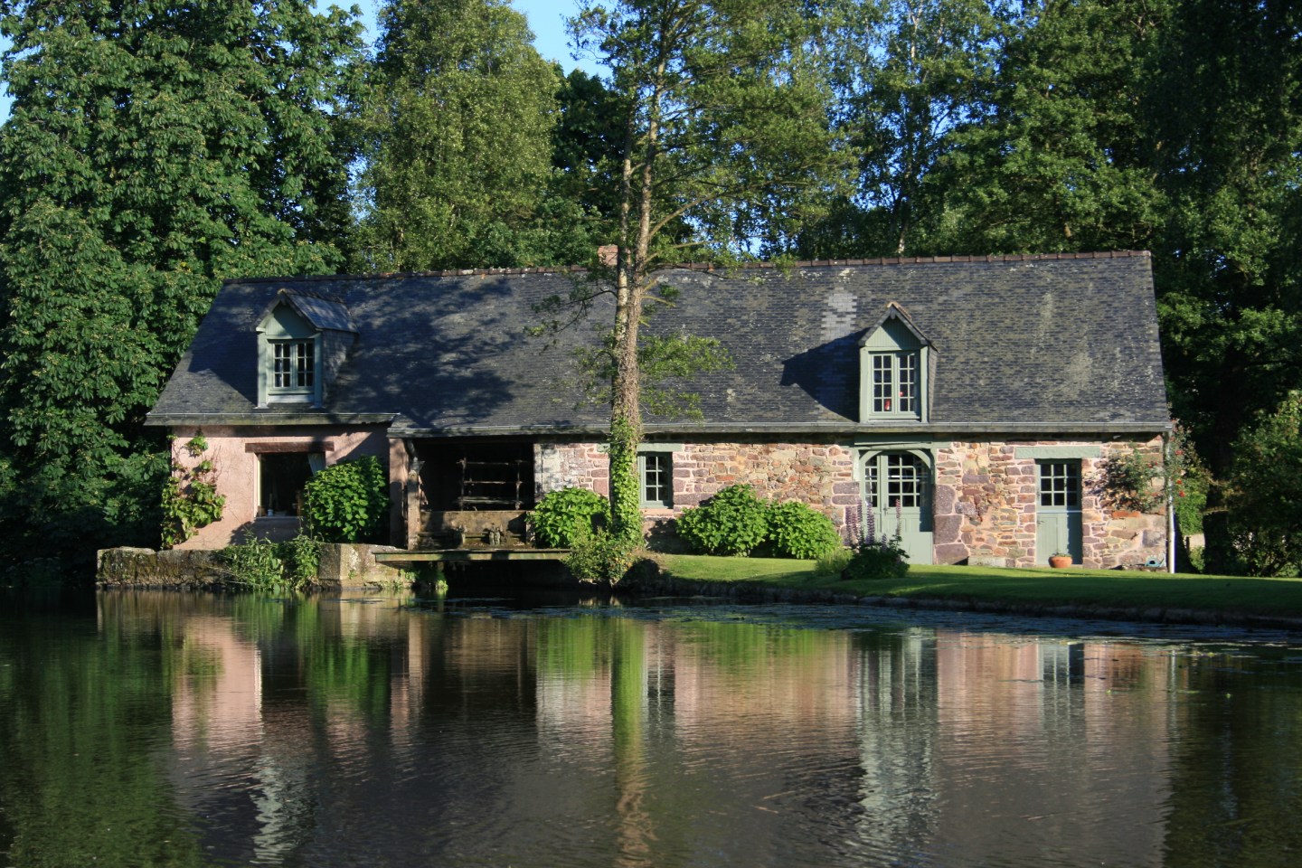 Ancien moulin en pierre et enduit rose au bord d'un étang avec reflet dans l'eau